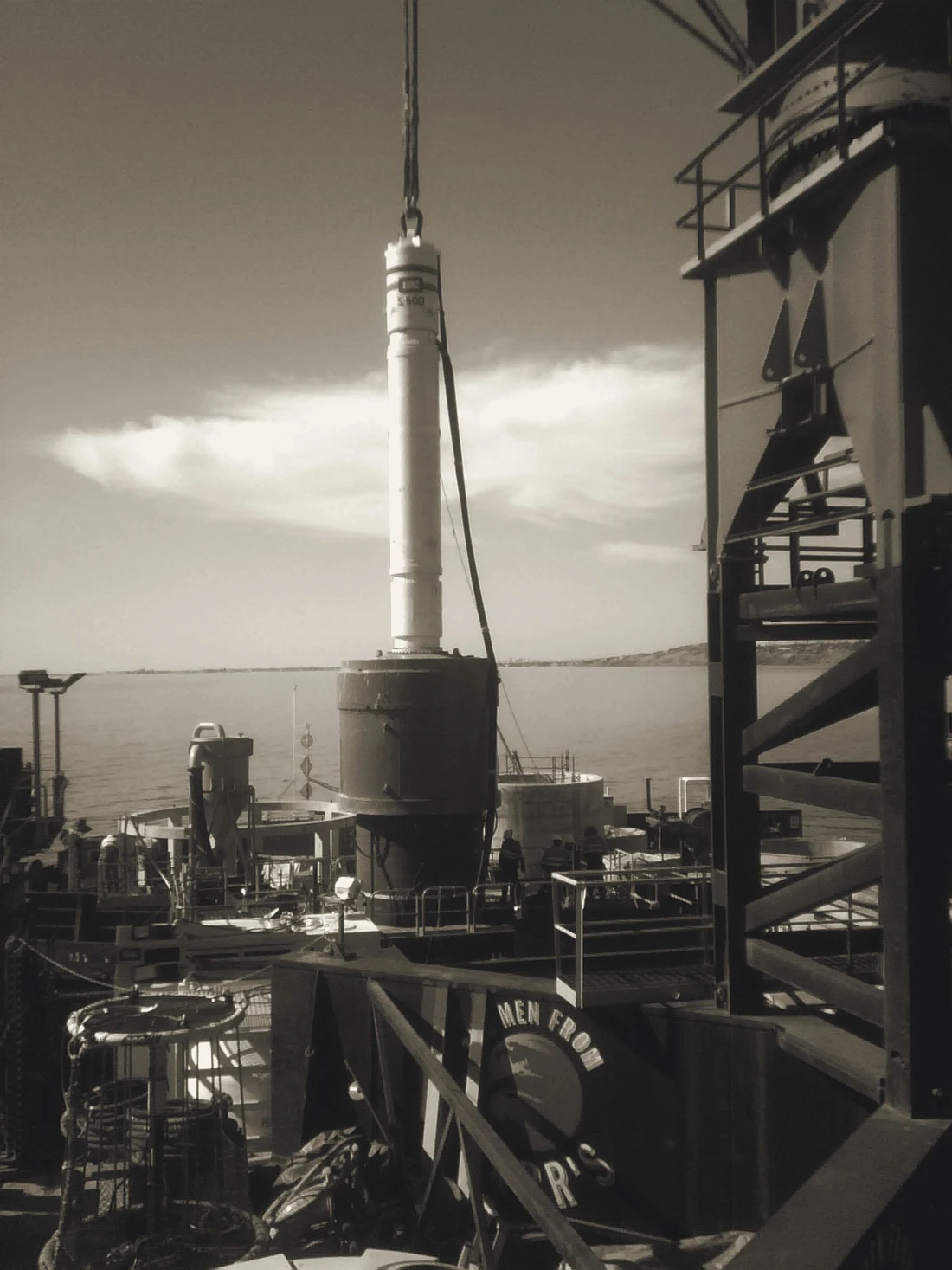 Large hydraulic piling hammer on a marine piling frame beside a coastal works vessel.